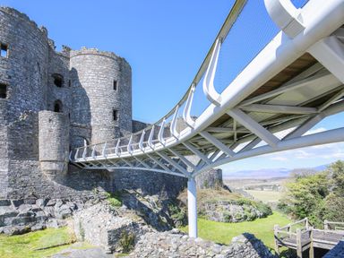 A stone castle with a modern white metal bridge leading to it surrounded by greenery and mountains in the background at Glyn Morfa in Harlech