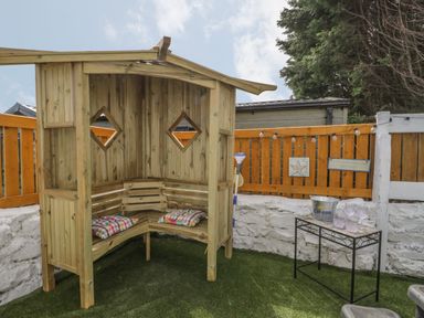 A wooden outdoor corner bench with two cushions on artificial grass next to a small table with glasses and a bucket outside at The Sequoia in Cloughton