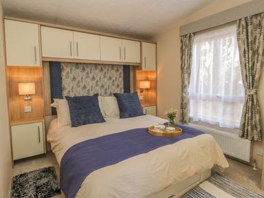 A bedroom with a double bed with blue cushions and a purple runner a tray with tea set on the bed bedside cabinets with lamps and a window with curtains at The Sequoia in Cloughton