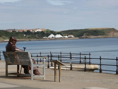 A metal statue of a person sitting on a bench by the waterfront with buildings and hills across the water