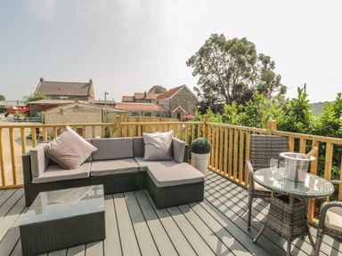 An outdoor deck with a gray sectional sofa a glass coffee table a round glass dining table with two chairs and a champagne bucket at The Maple in Cloughton