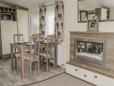 A dining area with a wooden table and four chairs set with plates and glasses next to a window with patterned curtains beside a built in electric fireplace in a modern room at The Maple in Cloughton