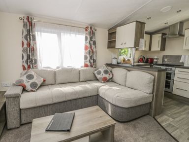 A living room with a beige and gray corner sofa, patterned curtains, a wooden coffee table, and a view of a kitchen area at The Maple in Cloughton