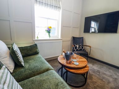 A living room with a green sofa, round wooden coffee tables, a black chair, a vase with flowers on the windowsill, and a wall-mounted television at Gavel Cottage in Whitby