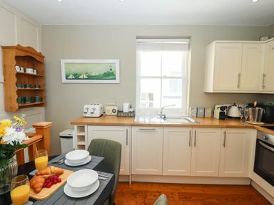 A kitchen with white cabinets a wooden countertop a sink a toaster a radio a table with two place settings croissants and orange juice and a wooden shelf with mugs at Gavel Cottage in Whitby
