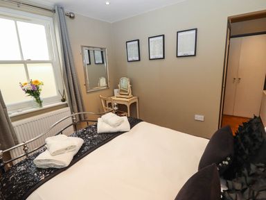 A bedroom with a bed, folded towels, a window with flowers, a dressing table with a mirror, and framed pictures on the wall at Gavel Cottage in Whitby