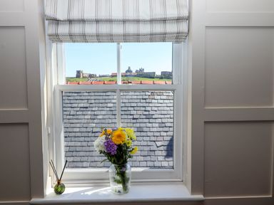 A window with a vase of flowers and reed diffuser on the sill overlooking rooftops and a distant building at Gavel Cottage in Whitby