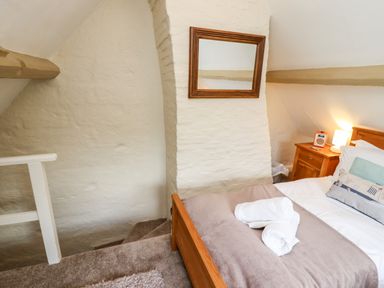 A bedroom with a bed, bedside table with a lamp and radio, stairs, and a wall mirror at Damson Cottage in Whitby