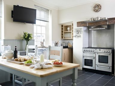 A kitchen with a wooden table set with bread fruit and wine next to a stainless steel stove and a wall mounted television at Albany House Whitby
