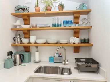 A kitchen corner with wooden shelves holding dishes and glasses above a stainless steel sink with a drying rack at The Cottage in Sandsend