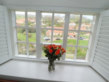 A vase with orange roses on a white windowsill with white shutters and view of houses and greenery outside at The Cottage in Sandsend