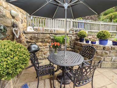 An outdoor patio area with a black metal table and four chairs under a large umbrella with plants and stone walls at The Cottage in Sandsend