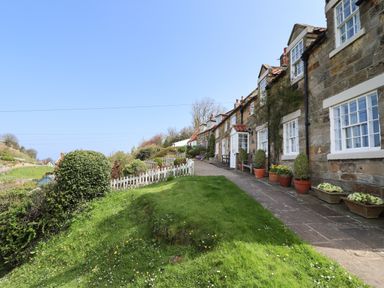 A row of stone cottages with white framed windows and potted plants outside along a pathway with a grassy area and white picket fence