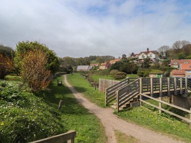 A dirt path with benches alongside and a wooden bridge crossing a small stream near houses and trees at The Cottage in Sandsend