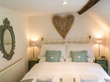 A bedroom with a bed with white bedding and green pillows two bedside tables with lamps a wicker heart wall decoration and a mirror at The Cottage in Sandsend
