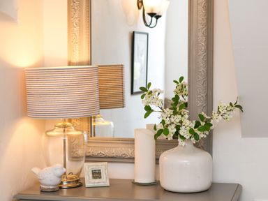 A tabletop with a lamp with striped shade a small bird figurine a framed photo a candle and a vase with white flowers in front of a mirror at The Cottage in Sandsend