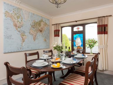 A dining room with a world map on the wall and a wooden table set with dishes glasses and bottles near a window with curtains and a stained glass panel