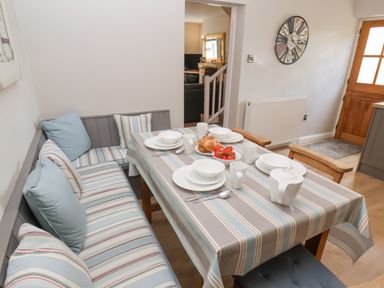 A dining area with a striped tablecloth on the table set with white dishes and mugs and a bench with striped cushions at Rooftops Cottage in Whitby