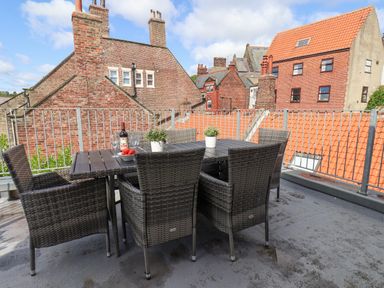 An outdoor patio with a dining table and wicker chairs with brick buildings in the background at Rooftops Cottage in Whitby
