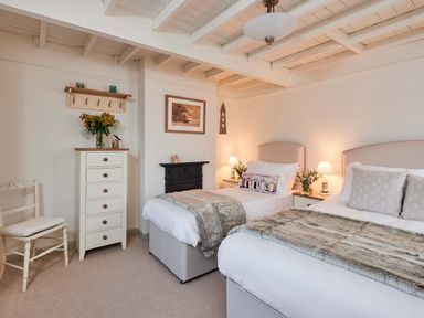 A bedroom with two beds with throw blankets and pillows bedside tables with lamps a chair a chest of drawers and a wall-mounted coat rack at Rigg Cottage Sandsend