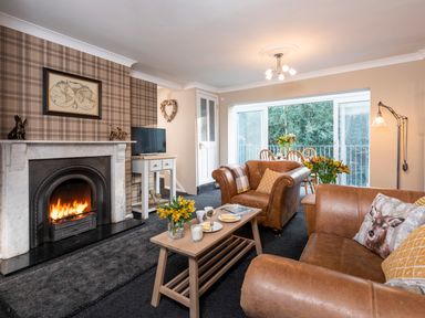 A living room with a fireplace two brown leather armchairs a wooden coffee table and a dining table by large windows at Esk View Cottage in Grosmont