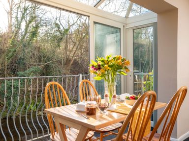 A dining area with a wooden table and chairs a vase of flowers and breakfast items by large windows overlooking trees at Esk View Cottage in Grosmont