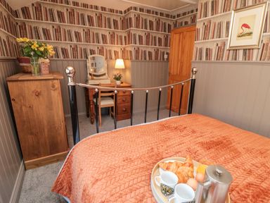 A bedroom with a metal bed frame a wooden dresser a desk with a chair and lamp and wallpaper with books at Esk View Cottage in Grosmont