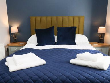 A bed with a mustard headboard blue pillows and blue quilted bedspread with white towels on top and bedside tables with lamps at Church View in Whitby