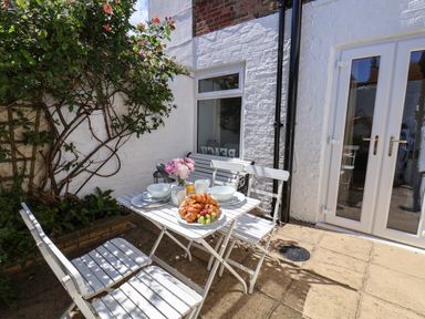 An outdoor patio with a white table set with croissants fruit and breakfast items surrounded by white chairs and a bench at Albany Cottage in Whitby