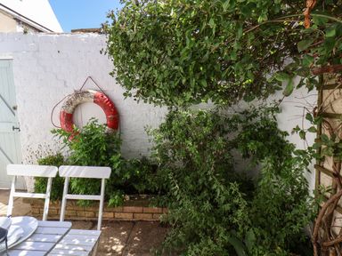 A garden patio with white chairs a red and white lifebuoy hanging on a white brick wall and green plants at Albany Cottage in Whitby