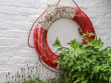 A worn red and white lifebuoy hanging on a white brick wall above green plants