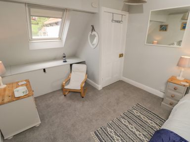 A bedroom with a window, a small chair, a dresser, bedside table with lamps and a patterned rug at Arden House Whitby