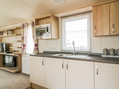 A kitchen area with a sink microwave cabinets and window next to a living room with shelves a television and curtains at The Hideaway in Cockermouth