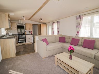 A living room with a beige sectional sofa and purple cushions next to a wooden coffee table with flowers in a vase and an open kitchen with wooden cabinets in the background at The Hideaway in Cockermouth