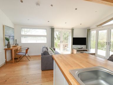 A living room with a grey sofa a wooden desk with a chair a wall-mounted television and large windows at 6 Valley View Lanreath