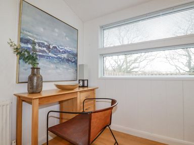 A small room with a wooden desk a chair a tall lamp a vase with greenery a bowl and a large painting above the desk at 6 Valley View in Lanreath