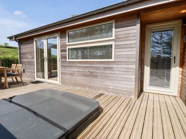 An outdoor wooden deck with a hot tub, wooden chairs, table, glass doors, and windows at 6 Valley View in Lanreath