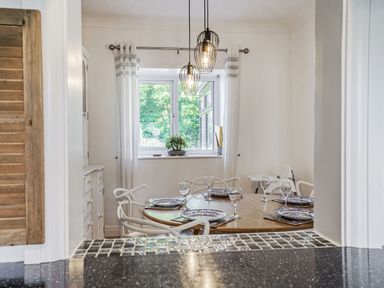 A dining room with a round wooden table set with plates and glasses white chairs a window with white curtains and hanging pendant lights at Granary Cottage in Pembroke