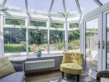 A conservatory with glass walls and ceiling showing garden outside with a yellow armchair a grey sofa a white side table and a plant on the windowsill at Granary Cottage in Pembroke