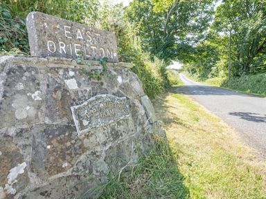 A country road with a stone sign saying East Orieston near trees and grass at Granary Cottage in Pembroke