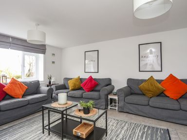 A living room with three gray sofas with colorful cushions and a glass coffee table at Gorwel in Moelfre