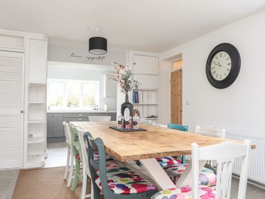 A dining room with a wooden table and multicolored chairs with a kitchen in the background at Gorwel in Moelfre