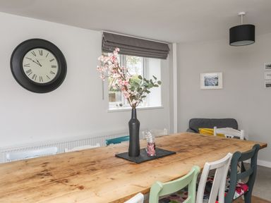 A dining room with a large wooden table surrounded by multicolored chairs a black vase with flowers on the table a black wall clock and a window with a gray shade at Gorwel in Moelfre