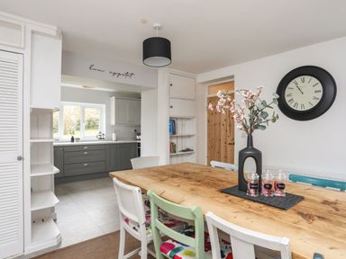 A dining room with a wooden table and chairs floral cushions and a black vase with flowers next to a kitchen with gray cabinets and a window at Gorwel in Moelfre