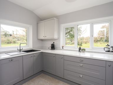 A kitchen with grey lower cabinets white upper cabinet a sink kettle and windows showing a garden at Gorwel in Moelfre