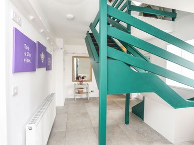 A hallway with green wooden staircase purple wall art a radiator and a small table with flowers and a mirror at Gorwel in Moelfre