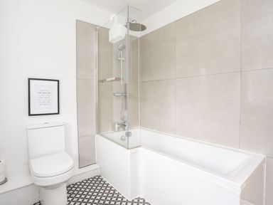 A bathroom with a toilet a bathtub with shower and glass panel and patterned floor tiles at Gorwel in Moelfre