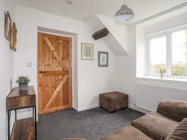 A small room with a wooden door a brown couch a matching ottoman a window a small table with a plant and framed pictures on the wall at Gorwel in Moelfre