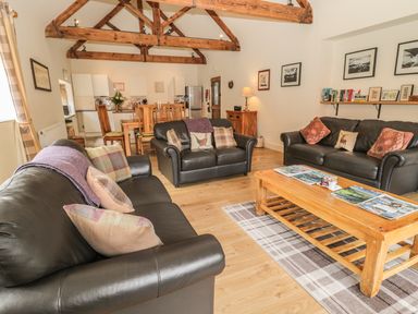 A living room with three black leather sofas a wooden coffee table with magazines and a wooden dining table in the background with exposed wooden ceiling beams at Sawmill Cottage in Belford near Bamburgh