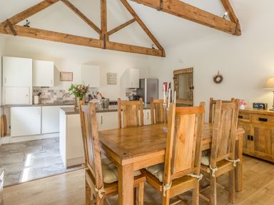 A dining area with wooden table and chairs next to a kitchen with white cabinets and wooden beams on the ceiling at Sawmill Cottage in Belford near Bamburgh